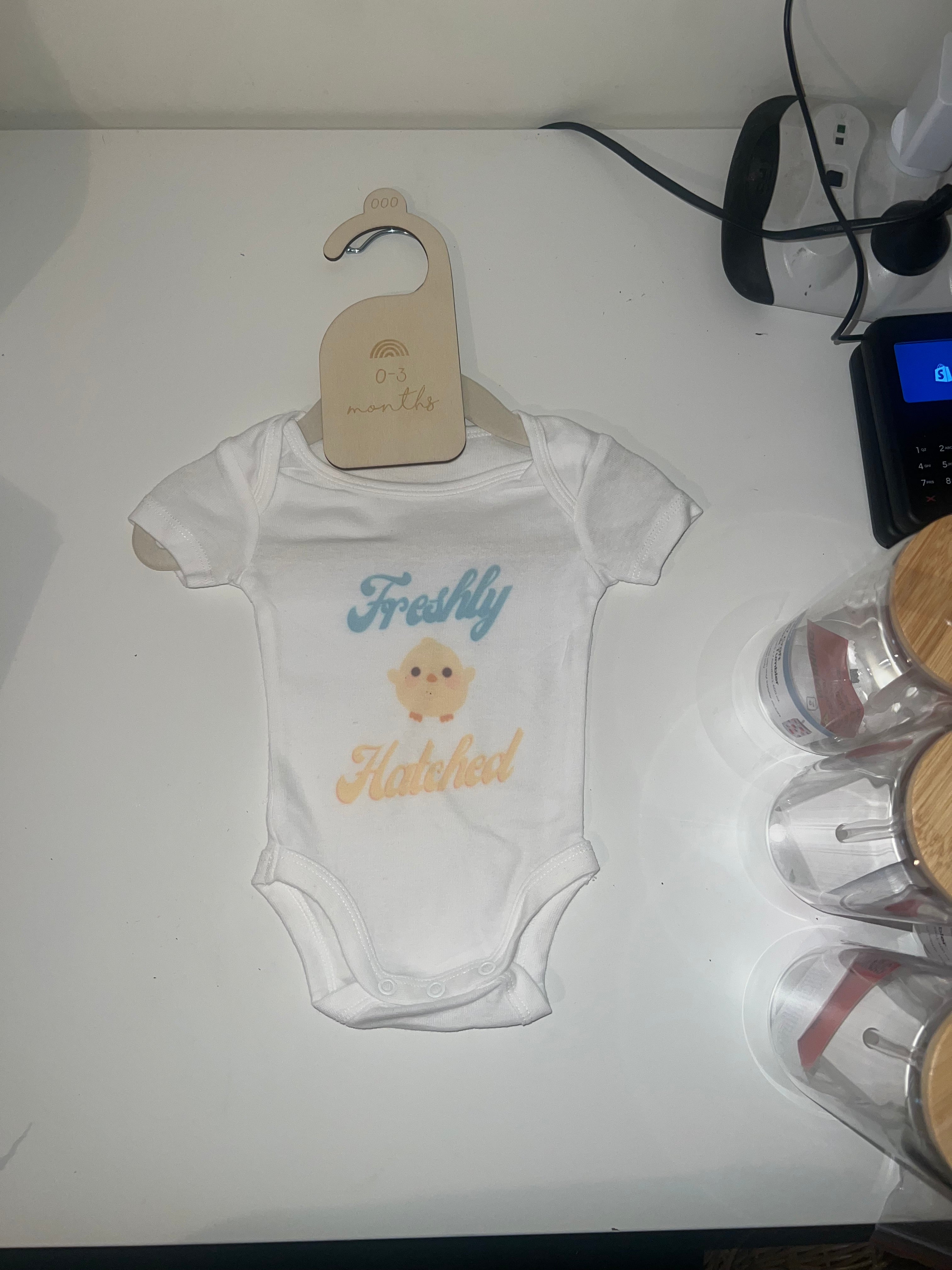 White short sleeve baby onesie with fluffy chick and Freshly Hatched text in blue and yellow on 0-3 months hanger
