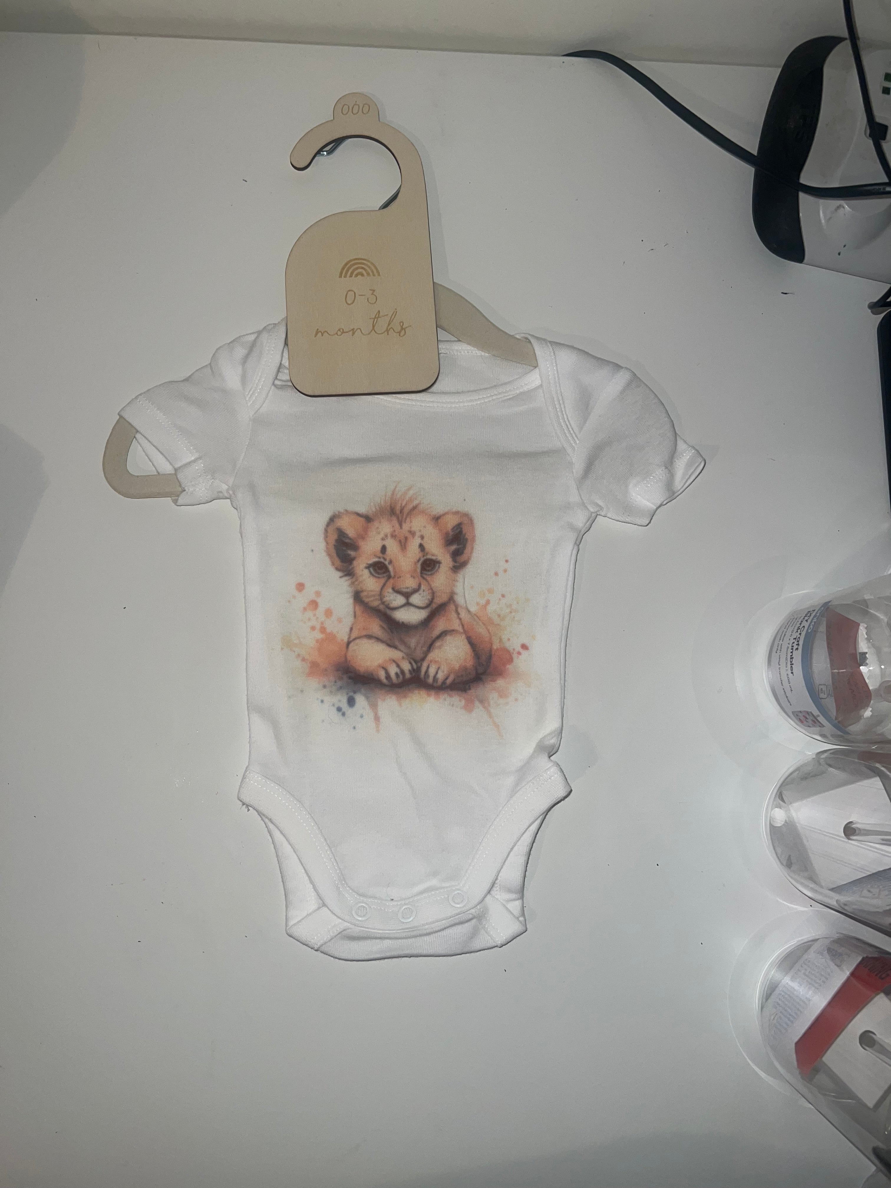 White short sleeve baby onesie with watercolour lion cub illustration in warm golden tones on 0-3 months hanger
