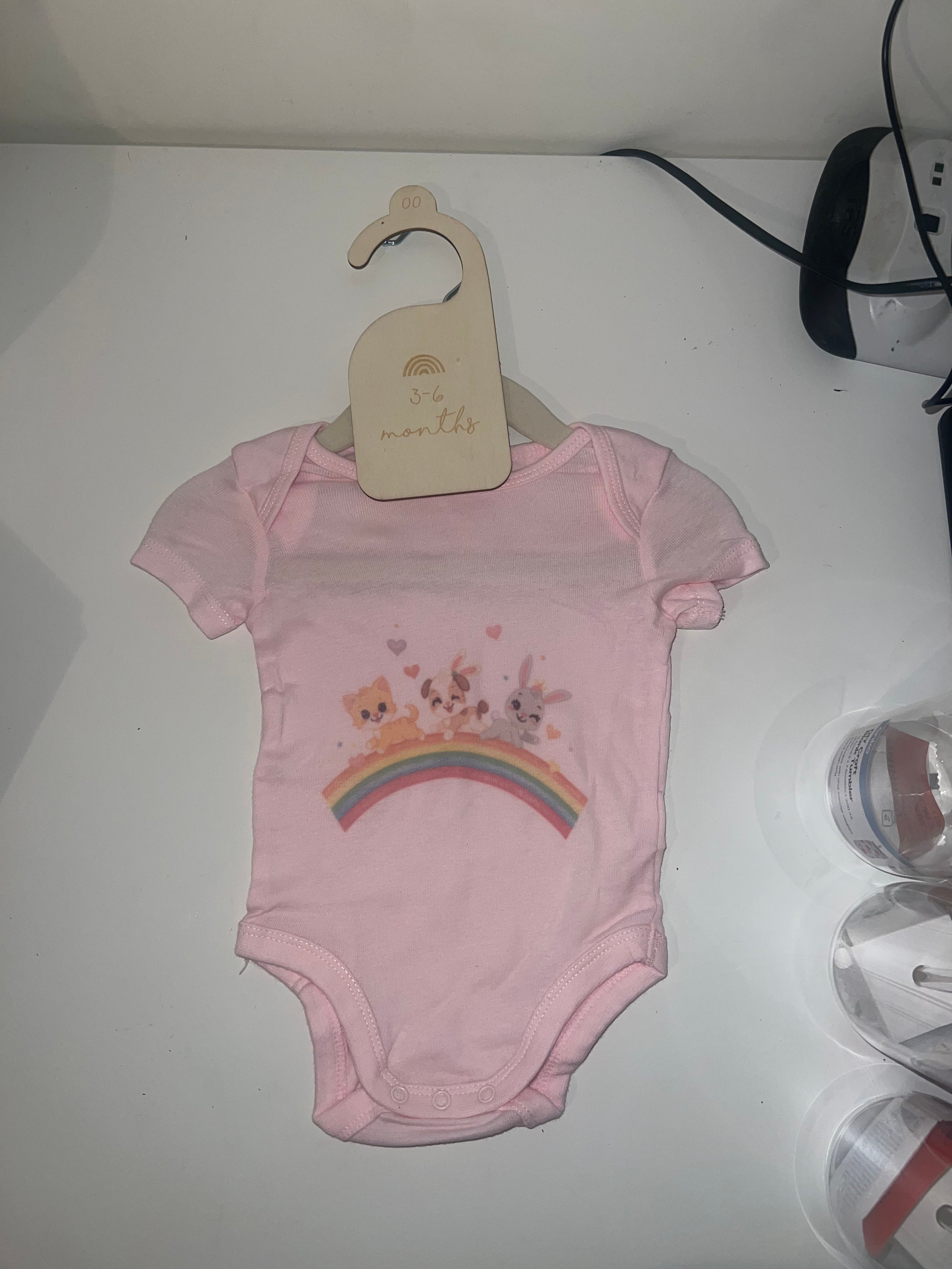 Blush pink short sleeve baby onesie with bear bunny and cat peeking over rainbow with hearts on 3-6 months hanger