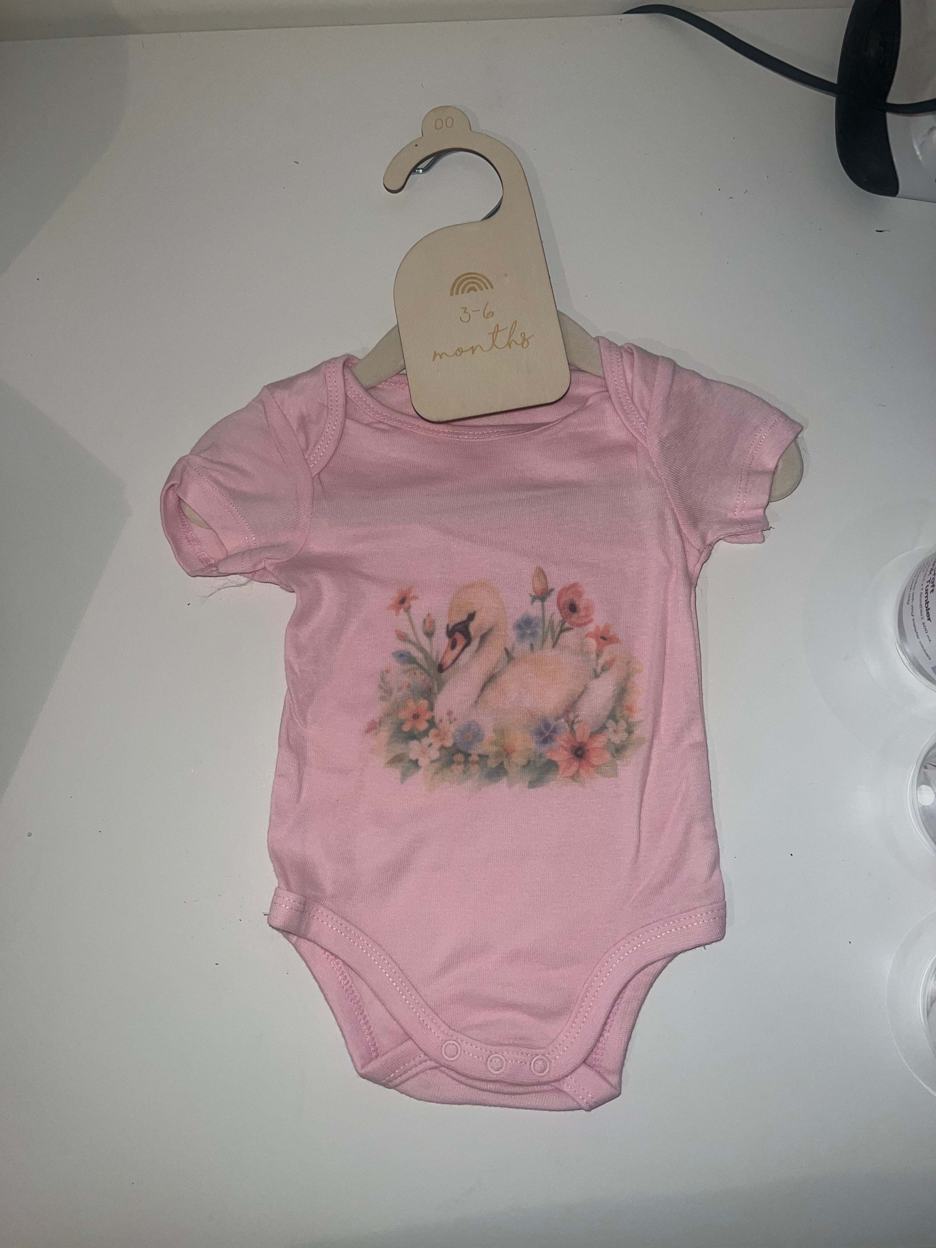 Soft pink short sleeve baby onesie with watercolour swan and pastel floral wreath on 3-6 months hanger
